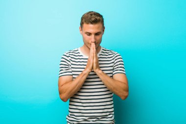 Genç beyaz adam ağzına yakın dua el ele mavi bir duvara karşı, kendinden emin hissediyor.