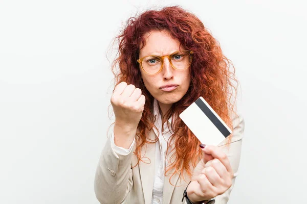 Young caucasian redhead woman holding a credit card showing fist to camera, aggressive facial expression.