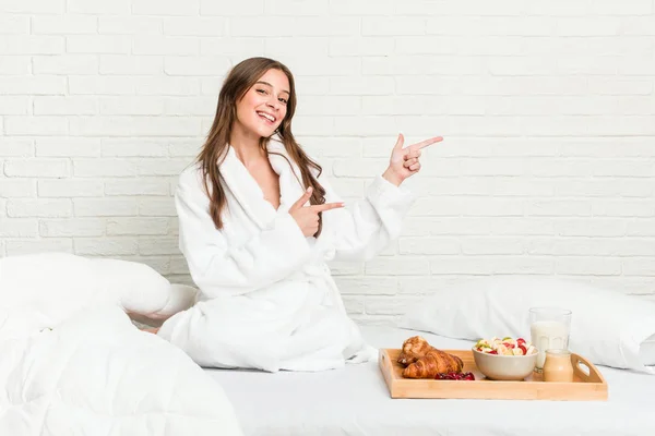 Young caucasian woman on the bed excited pointing with forefingers away.