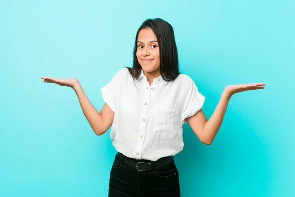 Young hispanic cool woman against a blue wall doubting and shrugging shoulders in questioning gesture.