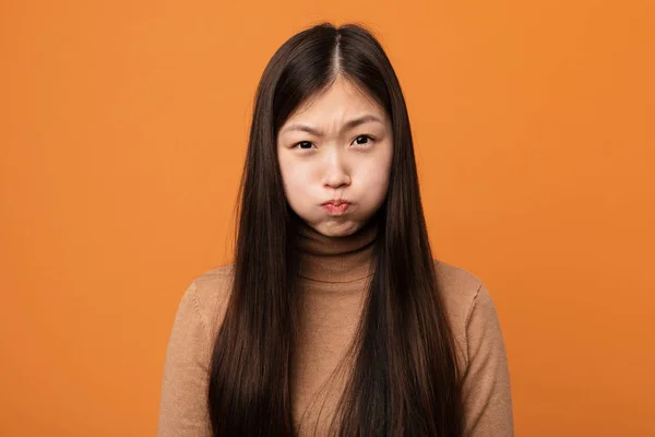 Young pretty chinese woman blows cheeks, has tired expression. Facial expression concept.