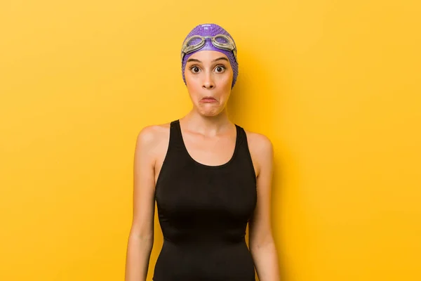 Young swimmer caucasian woman shrugs shoulders and open eyes confused.