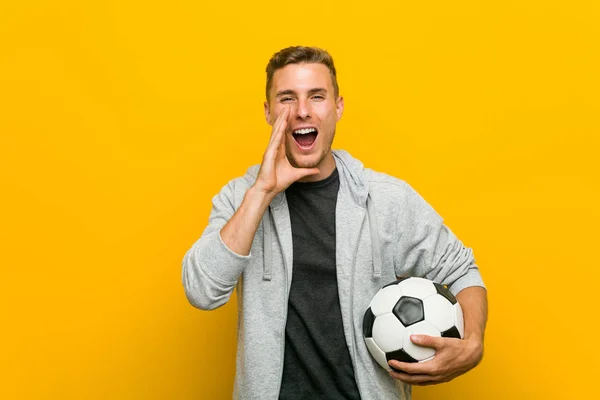 Młody Kaukaski Mężczyzna Trzyma Piłkę Nożną Krzycząc Podekscytowany Przodu — Zdjęcie stockowe