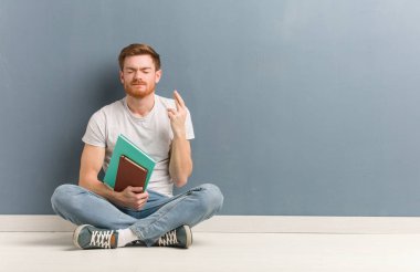 Genç kızıl saçlı öğrenci adam şans için parmaklarını geçerek yerde oturuyor. Elinde kitap var..