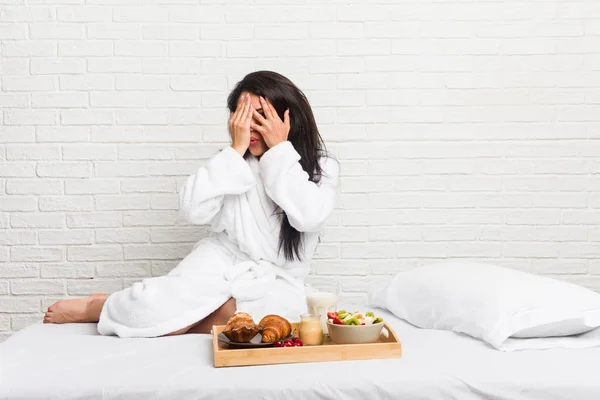 Young curvy woman taking a breakfast on the bed blink through fingers frightened and nervous.
