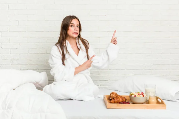 Young caucasian woman on the bed shocked pointing with index fingers to a copy space.