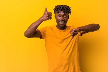 Genç siyah adam sarı arka plan üzerinde başparmak yukarı ve başparmak aşağı gösteren rastas giyen, zor kavramı seçin