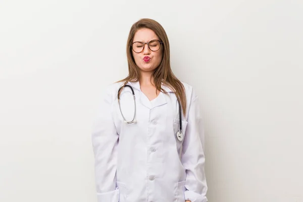 Young doctor woman against a white wall blows cheeks, has tired expression. Facial expression concept.