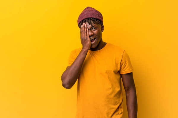 Young black man wearing rastas over yellow background having fun covering half of face with palm.
