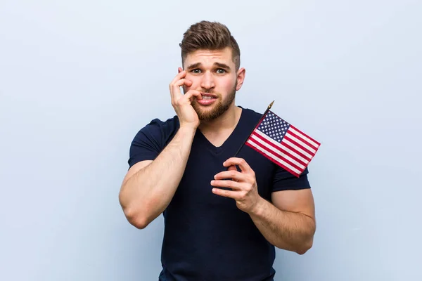 Young caucasian man holding a united states flag biting fingernails, nervous and very anxious.