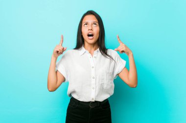 Young hispanic cool woman against a blue wall pointing upside with opened mouth.