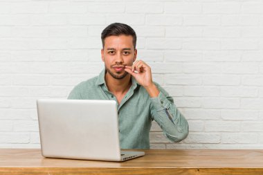 Genç filipinli adam dudaklarında parmakları ile dizüstü bilgisayar ile çalışan bir sır tutmak oturuyor.