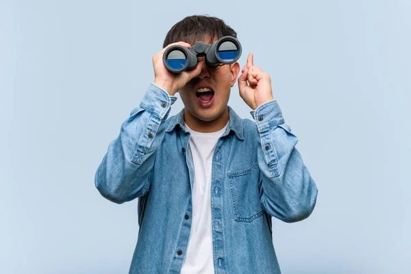 Young chinese man holding a binoculars crossing fingers for having luck