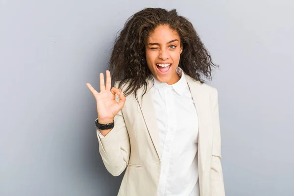 Young african american business woman winks an eye and holds an okay gesture with hand.