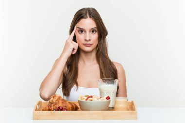 Young caucasian woman having breakfast pointing his temple with finger, thinking, focused on a task.