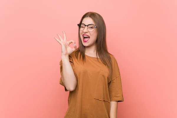 Young student woman wearing eyeglasses winks an eye and holds an okay gesture with hand.