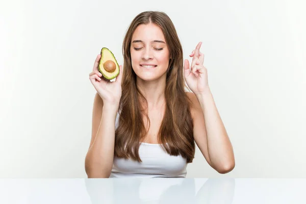 Young Caucasian Woman Holding Avocado Crossing Fingers Having Luck — Zdjęcie stockowe