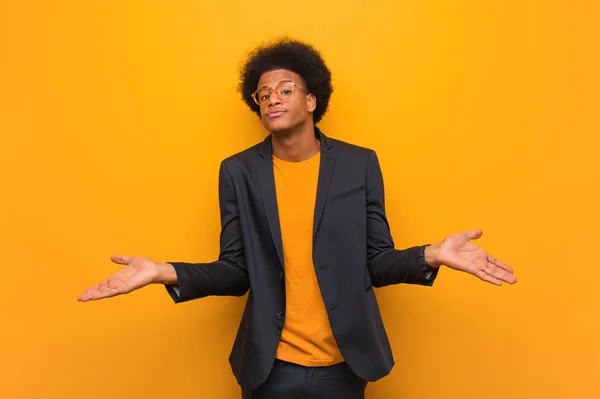 Young business african american man over an orange wall doubting and shrugging shoulders