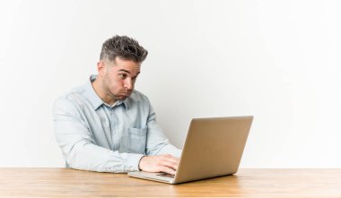 Young handsome man working with his laptop blows cheeks, has tired expression. Facial expression concept.