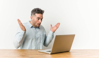 Young handsome man working with his laptop surprised and shocked.