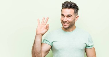 Yeşil bir arka plan karşı Genç yakışıklı adam bir göz kırpmave el ile tamam bir jest tutar.