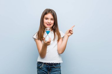 Küçük beyaz kız bir kum saati tutan neşeyle işaret parmağı ile işaret gülümseyerek uzakta.