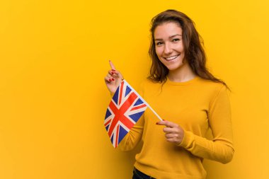Birleşik krallık bayrağı tutan genç Avrupalı kadın neşeyle işaret parmağıyla işaret ederek gülümsüyor.