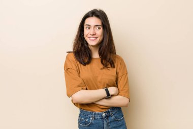 Genç esmer kadın bej bir arkaplanda kollarını kavuşturarak gülümsüyor..