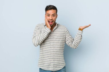 Genç yakışıklı filipinli adam bir avuç üzerinde kopya alanı tutar, yanak üzerinde el tutmak. Şaşırmış ve memnun.