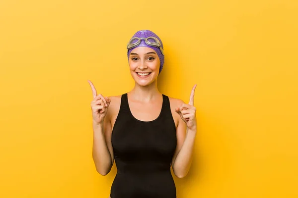 Young swimmer caucasian woman indicates with both fore fingers up showing a blank space.