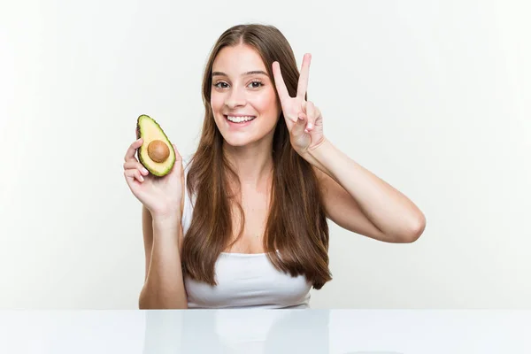 Joven Mujer Caucásica Sosteniendo Aguacate Mostrando Signo Victoria Sonriendo Ampliamente — Foto de Stock