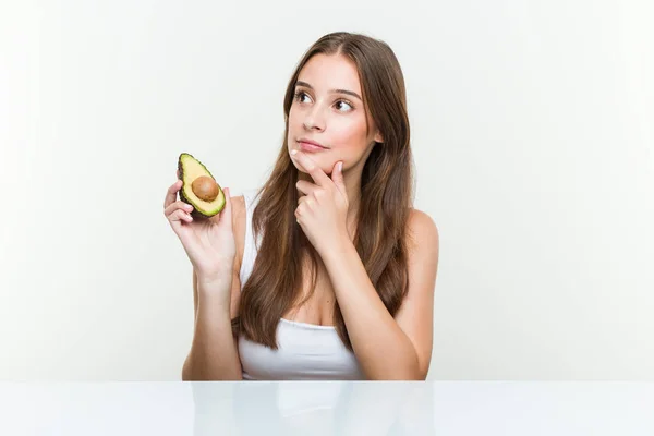 Joven Mujer Caucásica Sosteniendo Aguacate Mirando Lado Con Expresión Dudosa —  Fotos de Stock