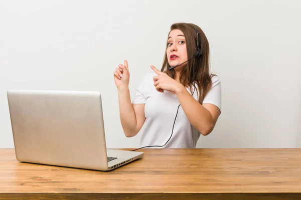 Young telemarketer woman shocked pointing with index fingers to a copy space.