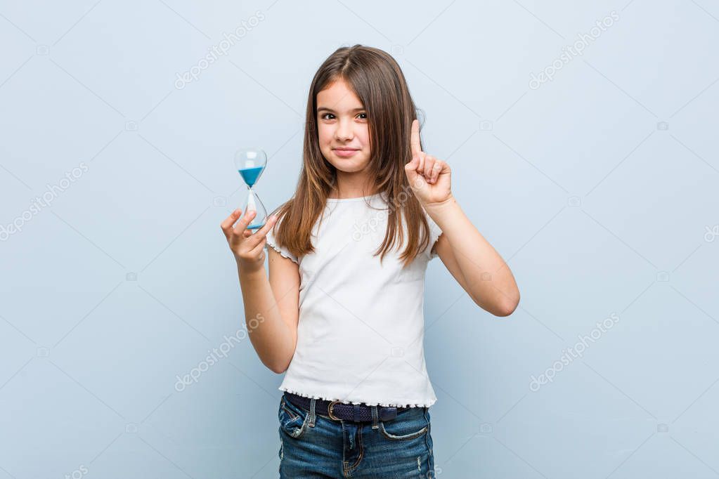 Little caucasian girl holding an hourglass showing number one with finger.