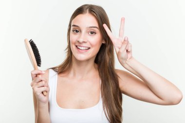 Genç beyaz kadın zafer işareti gösteren bir saç fırçası tutan ve geniş gülümseyen.