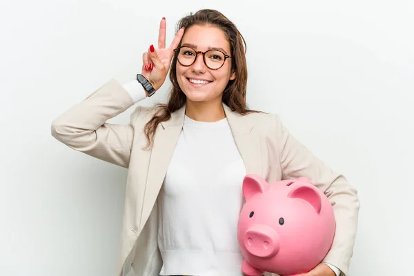 年轻的欧洲女商人拿着一个小猪银行 显示胜利的标志和微笑广泛 — 图库照片