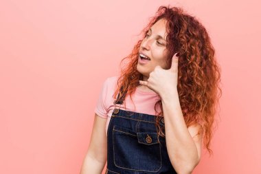 Genç güzel kızıl kızıl saçlı kadın parmakları ile bir cep telefonu arama jest gösteren bir kot dungaree giyiyor.