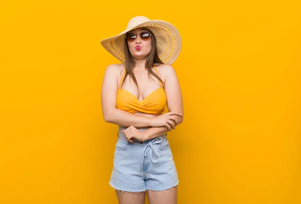 Young caucasian woman wearing a straw hat, summer look blows cheeks, has tired expression. Facial expression concept.