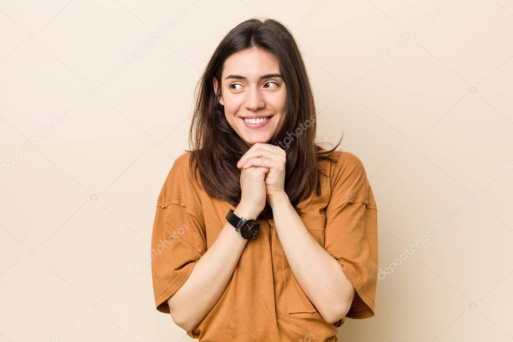 Young brunette woman against a beige background keeps hands under chin, is looking happily aside.
