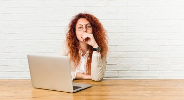 Genç kızıl saçlı kıvırcık kadın dizüstü bilgisayar ile çalışan mutlu ve kendinden emin gülümseyerek, el ile çene dokunmadan.