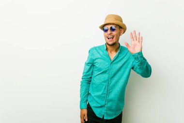 Yazlık kıyafetler giyen genç İspanyol adam gözünü kırpıyor ve eliyle iyi bir jest yapıyor..
