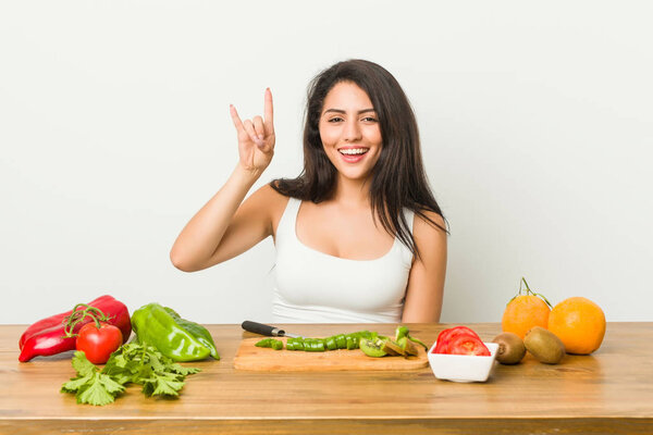 Молодая пышная женщина готовит здоровую еду, показывая жест рога, как концепция революции
.