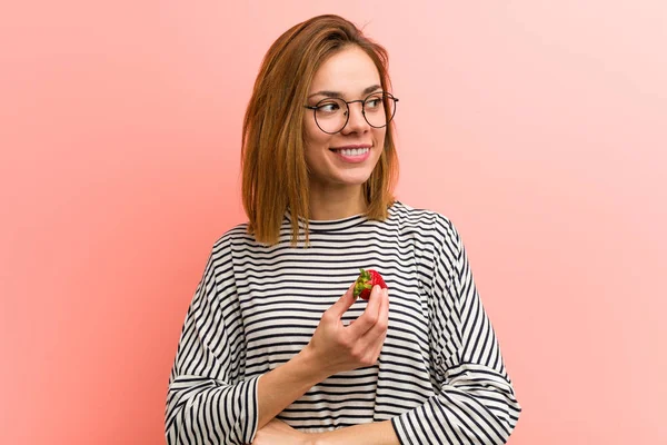 Young woman holding a strawberry smiling confident with crossed arms.