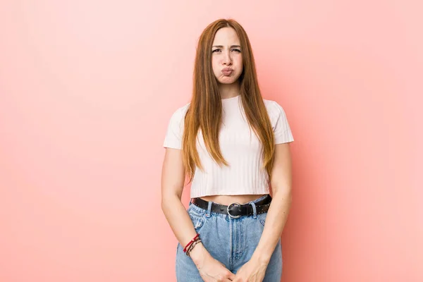 Young redhead ginger woman against a pink wall blows cheeks, has tired expression. Facial expression concept.