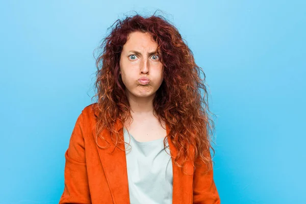 Young redhead elegant woman blows cheeks, has tired expression. Facial expression concept.