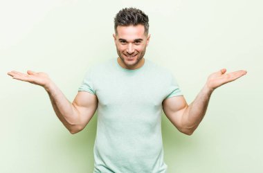 Yeşil bir arka plan karşı Genç yakışıklı adam kolları ile ölçek yapar, mutlu ve kendinden emin hissediyor.