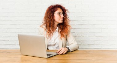 Dizüstü bilgisayarıyla çalışan genç kızıl saçlı kıvırcık kadın gülümseyen, neşeli ve hoş bir kenara bakıyor.