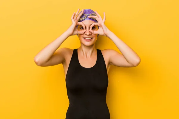 Young swimmer caucasian woman showing okay sign over eyes