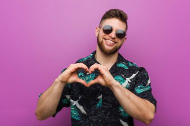 Tatil eki beslenen genç adam gülümseyerek ve elleriyle kalp şekli gösteriyor..