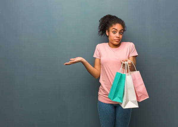 Young black woman doubting and shrugging shoulders. She is holding a shopping bags.